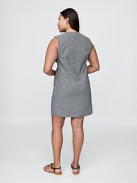 Femme se tenant debout, les bras fléchis, portant une robe sans manches en vichy noir et blanc et des sandales plates noires sur un fond uni.