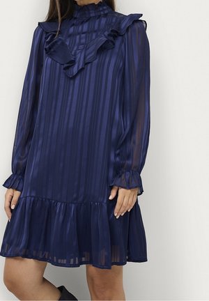 Femme portant une robe à rayures bleu marine transparente avec un col, des poignets et un ourlet volantés, debout devant un fond clair uni.