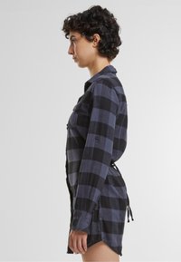 Abito camicia in flanella a quadri blu e neri con maniche lunghe, polsini con bottoni, due taschini sul petto e vita regolabile con cordoncino.