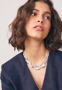 Collier en argent avec des maillons ovales et en forme de croissant, associé à un haut à col en V de couleur bleu marine foncé. La surface lisse et la finition brillante mettent en valeur son design.