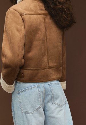 Personne aux cheveux ondulés portant une veste en daim marron avec des poignets en peau retournée et un jean taille haute bleu clair, vue de dos.