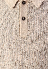 Μπεζ πλεκτό πουκάμισο πόλο με υφή, παραδοσιακό γιακά και τρία μαύρα κουμπιά. Διακριτό στίγμα ανάγλυφου σχεδίου σε όλη την επιφάνεια.