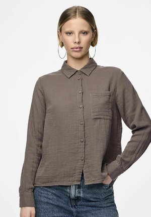 Camisa marrón de botones hecha de una tela texturada con cuello, un bolsillo en el pecho y dobladillo ligeramente recortado. El modelo lleva jeans de mezclilla.