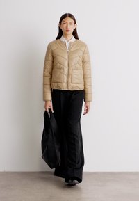Beige quiltet pufferjakke med chevron mønster, frontlynlås, to lommer; modellen er sat sammen med sorte vide bukser og en sort taske.