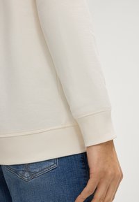 Jasno beżowa, długoskrzydła bluzka z prążkowanymi mankietami i dołem, o fakturowanym materiale, zestawiona z niebieskimi dżinsami z charakterystycznym wzorem kieszeni.