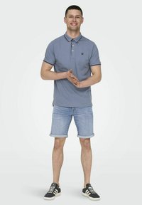 Camisa polo de color azul claro con cuello y mangas cortas, combinada con pantalones cortos de denim arremangados y zapatillas negras con detalles blancos.