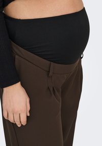 Braune Hose mit maßgeschneidertem Design, versehen mit Falten und einer Seitentasche. Kombiniert mit einem schwarzen Stretch-Oberteil, das eine runde Bauchform betont.