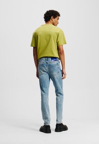 Tricou verde cu mânecă scurtă, asociat cu blugi denim de culoare albastră deschisă, având un patch cu logo pe talie, purtat cu pantofi negri.