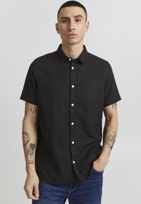 Kortærmet sort skjorte med knapper i linned, der har en brystlomme og hvide knapper, båret med blå jeans.