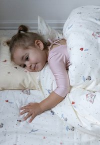 Dziecko odpoczywa na białym łóżku z wzorzystą pościelą, na której znajdują się kolorowe ilustracje. Jasnoróżowa bluzka z długim rękawem, miękkie poduszki, przytulna pościel.