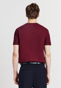 Camicia corta bordeaux con tessuto texture. Presenta un collo tondo e un taglio dritto, infilata in pantaloni blu navy con una cintura nera.