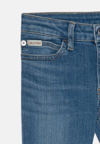 Jeans de mezclilla azul, diseño de cinco bolsillos con una cintura de altura media. Costuras a contraste, herrajes plateados y una etiqueta roja y verde en el bolsillo delantero.