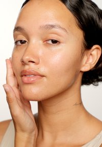 Nærbillede af et ansigt med glat, strålende hud; lys brun hudfarve, naturlig makeup og subtile fregner. En hånd berører blidt kinden.