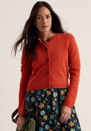 Mujer con el pelo largo y oscuro lleva un cárdigan naranja abotonado sobre un vestido azul marino de flores, sostiene un bolso verde y está de pie contra un fondo claro.
