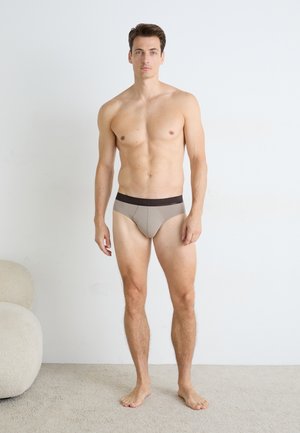 Beige Herrenunterwäsche mit einem dunklen Taillenbund, die ein eng anliegendes Design und eine glatte Textur aufweist. Das Modell steht barfuß vor einem neutralen Hintergrund.
