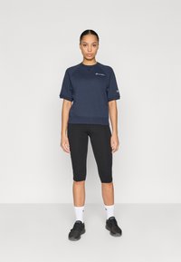 Μπλε σκούρο φούτερ με κοντά μανίκια, ριμπ τελείωμα, κεντημένο λογότυπο, συνδυασμένο με μαύρο κολάν μέχρι το γόνατο και μαύρα αθλητικά παπούτσια. Ορατές λευκές κάλτσες.