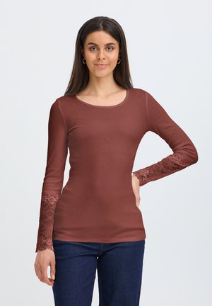 Jeune femme aux longs cheveux foncés porte un haut marron ajusté avec des manches en dentelle et un jean bleu foncé, debout devant un fond clair uni.