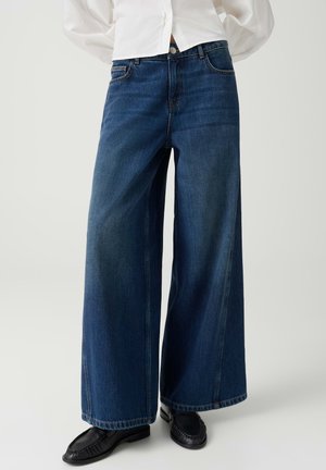 Femme portant un jean large bleu foncé associé à des mocassins noirs et une chemise blanche, montrée de la taille aux pieds.