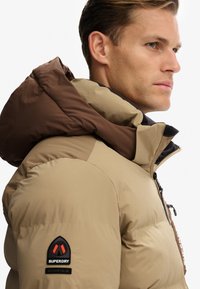 Tan pufferjakke med brun hætte, lynlåslukning og et mærket patch på skulderen. Glat tekstur og isoleret design for varme.