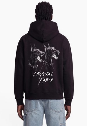 Man wearing black hoodie with white graphic of two snarling dogs and text "Crystal Paris" on the back, paired with light blue jeans.