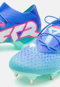 Scarpe da calcio blu con tomaia in mesh testurizzato, dettagli rosa e bianchi, e una suola traslucida con tacchetti metallici.