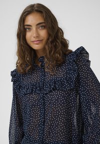 Fiatal nő, hullámos barna hajjal, sötétkék áttetsző blúzban, fehér pöttyökkel és fodros részletekkel a vállán és a mellkasán.