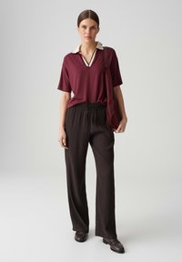 Polo bordeaux avec un col blanc, associé à un pantalon large plissé noir. Le mannequin tient un sac assorti et porte des chaussures foncées.