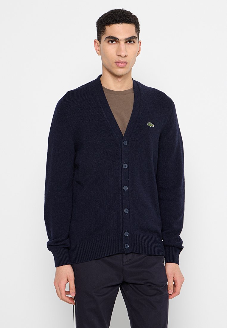 Cardigan lavorato a maglia blu navy con scollo a V, chiusura a bottoni e orlo a coste. Presenta un piccolo logo verde sul lato sinistro.