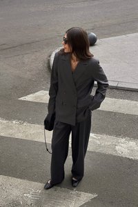 Šedý dvojradový blazer a voľné nohavice, doplnené čiernymi plochými topánkami. Doplnky zahŕňajú malú čiernu tašku a slnečné okuliare.