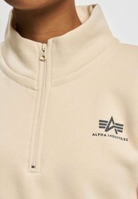 Beige halv-zip genser laget av mykt stoff. Har en høy krage og en svart logo med stiliserte bokstaver på venstre framside.