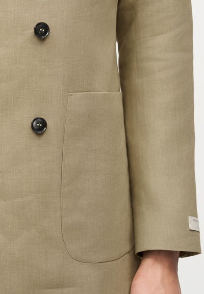 Μανίκι και μπροστινό μέρος μπεζ λινής ζακέτας με δύο μαύρα κουμπιά και μπροστινή τσέπη με ραφή, που δείχνει μια μικρή λευκή ετικέτα της μάρκας στο μανίκι.