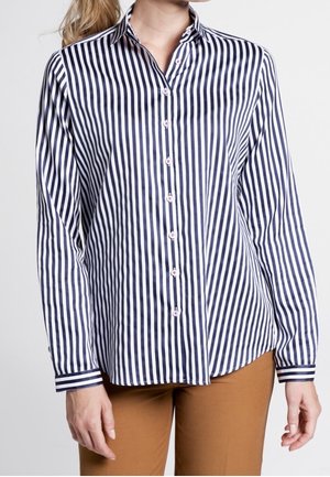 Vrouw draagt een marineblauw-wit verticaal gestreept overhemd met knopen en lange mouwen en beige broek, staand voor een effen achtergrond.