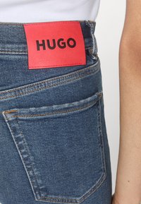 Jean en denim bleu avec une étiquette en cuir rouge arborant le mot "HUGO" en noir. Couture visible et détail de poche arrière.