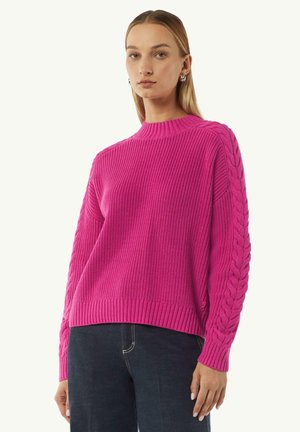 Pink knitted sweater with a ribbed texture and cable knit accents on the sleeves. Round neck and cropped fit, paired with dark jeans.