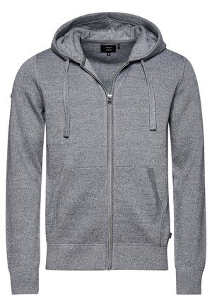 Mand iført grå hættetrøje med lynlås, hvid t-shirt, mørke bukser og hvide sneakers, stående med den ene hånd i lommen mod en ensfarvet baggrund.