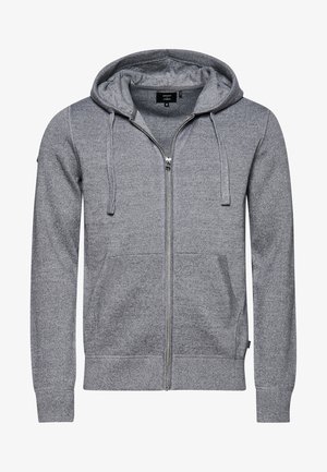 Mann trägt grauen Zip-up-Hoodie, weißes T-Shirt, dunkle Hose und weiße Sneakers, steht mit einer Hand in der Tasche vor einfarbigem Hintergrund.