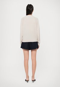 Maglione beige chiaro con maniche lunghe, colletto a giro e orlo a coste, abbinato a pantaloni corti blu navy. Texture liscia, design semplice, senza motivi visibili.