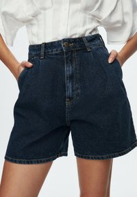 Riflové šortky tmavě modré barvy s vysokým pasem, mají přední zapínání na knoflíky, poutka na opasek a boční kapsy. Texturovaná látka s viditelným prošitím.