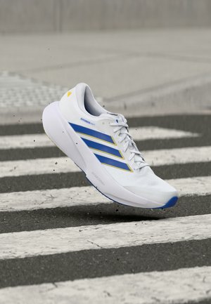 Λευκό αθλητικό παπούτσι με μπλε και κίτρινες ρίγες, στον αέρα πάνω από διάβαση πεζών σε ασφαλτόδρομο, με σωματίδια βρωμιάς γύρω από την σόλα.