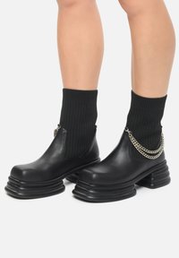 Svarta ankelskor med en kraftig plattform, överdel i släta läder, ribbade svarta strumpor och en dekorativ silverkedjeaccent.