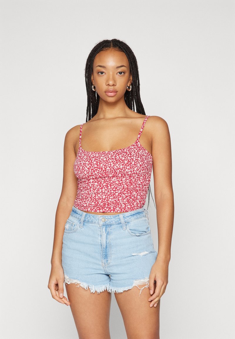 Červený kvetinový crop top s tenkými ramienkami, ktorý má smockovaný dizajn a vzory bielych kvetov, skombinovaný s svetlomodrými roztrhnutými džínsami.