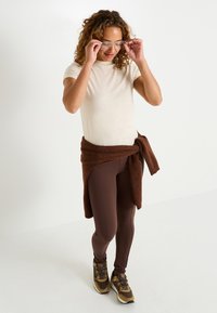 Camiseta de manga corta beige, leggins marrones y un suéter marrón atado a la cintura. Zapatillas deportivas marrones y doradas con un acabado texturizado.