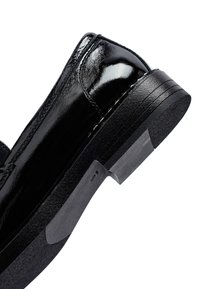 Zapato de vestir de cuero negro brillante con un tacón de goma bajo y apilado, y un acabado suave y pulido sobre un fondo blanco.