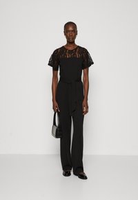 Combinaison noire avec des manches courtes en dentelle, une taille ceinturée, un pantalon large et un sac à main texturé. Tissu lisse avec une silhouette élégante.