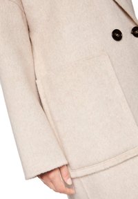 Beige wool coat with a soft texture, featuring a large front pocket and two dark buttons on the double-breasted design.