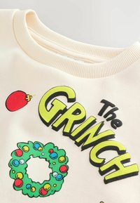 Krémový sweatshir s tučným zeleným nápisom "Grinch", s farebným vencom a červenou ozdobou v grafike. Textúrovaný ribbovaný výstrih.