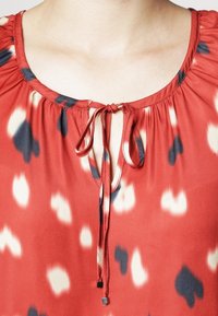 Blusa rossa con collo rotondo, dettaglio arricciato e chiusura con lacci. Presenta un motivo astratto colorato con accenti blu navy e crema.