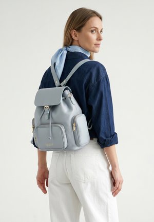 Vrouw met een lichtblauwe sjaal geknoopt om de nek draagt een marineblauw overhemd, witte broek en draagt een lichtblauwe leren rugzak met het label "VENEZIA."