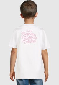 Camiseta de algodón blanca con texto en cursiva rosa en la parte posterior que dice "Abraza Cada Momento". Cuello redondo y mangas cortas.