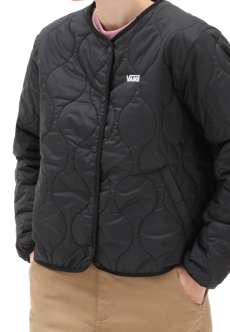 Personne portant une veste matelassée noire Vans avec des boutons-pression, deux poches avant et un pantalon beige, mains dans les poches.